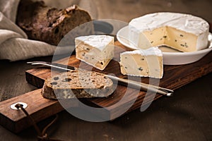 Loaf of soft blue cheese from cow milk on porcelain plate with walnut bread, knife, linen towel and dark brown wooden board as