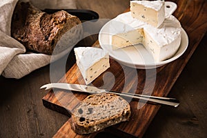 Loaf of soft blue cheese from cow milk on porcelain plate with walnut bread, knife, linen towel and dark brown wooden board as