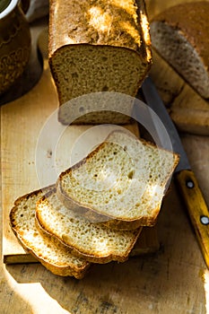 Loaf of sourdough bread cut into slices on wood cutting board, knife, kitchen table, sunlight flecks, cozy