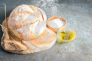 Loaf of white round bread, some wheat ears, white flour and olive oil on the dark background. Natural handmade white bread