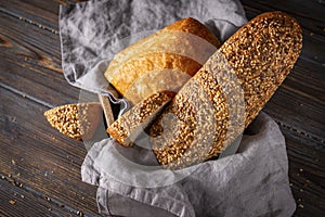 Loaves and slices of fresh grain bread in a basket with a towel on a dark wooden background