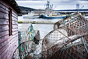 Lobster traps and crab traps stacked on a dock with fishing trawlers at background at Whiteway Newfoundland