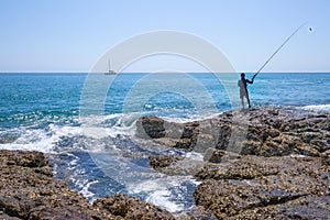 A local fisher man with his fishing rod is looking the boat in the ocean on rocks sea coast, shore of the Bay in Asia, Thailand