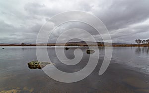 Loch Ba on Rannoch Moor in the Highlands of Scotland on a wet overcast day in April, with boulders on the surface of the water.