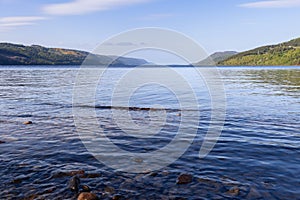 Loch Ness presents a serene view under clear skies, with its vast water expanse bordered by sloping green hills