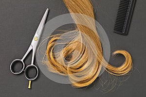 A lock of blond curly female hair, scissors and a comb on a dark background, top view