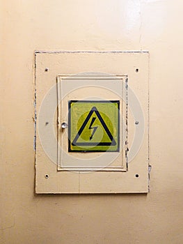Locked electrical panel on the wall painted with yellow oil paint. There is a high voltage sign on the door