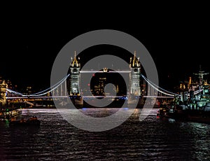 London bus driving past Tower Bridge illuminated at night with illuminated skyscrapers and the war museum ship HMS Belfast docked