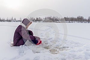 A lone angler girl is concentrating on catching fish with a winter fishing rod sitting on a substrate on the ice among flakes