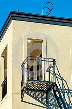 Lone balcony with fire escape ladder and rooftop access with beige stucco exterior with blue sky clear background