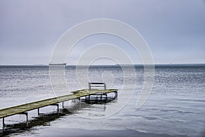 Lone bench on a thin boardwalk at sea and a faraway vessel amid winter gloominess covey sadness, solitude and desolation feelings