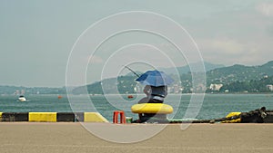 A lone fisherman sits on the city's waterfront. A man with a fishing rod is hiding from the sun under an umbrella