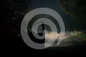 A lone hooded figure standing on an empty misty spooky road at night. Highlighted with car headlights.