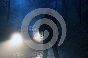 A lone hooded figure standing in front of car headlights in the middle of a country road on a spooky, eerie, moody foggy night