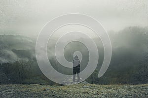 A lone hooded figure on top of a hill, looking out across the countryside on a foggy, rainy winters day.