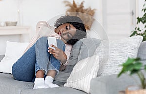 Loneliness. Depressed Black Lady Sitting On Couch With Smartphone, Loking At Screen