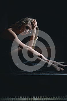 Sad beautiful girl sitting on black piano in the dark room