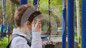 Lonely beautiful kid wear white shirt, protective medical mask, ride on swing with rusty chains on blurred background