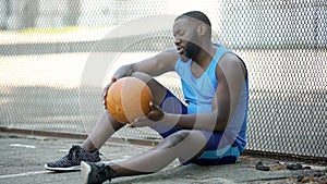 Lonely black basketball player sitting on stadium ground and holding ball, sport