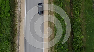 Lonely black car drives on asphalt road going through the green forest. Aerial view