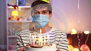 Lonely Caucasian young man in Covid face mask and party hat holding birthday cake and looking at camera. Portrait of sad
