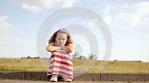 Lonely curly girl sitting on the edge of the road against the blue sky. The girl is sad and angry