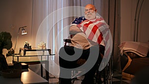 A lonely elderly disabled man wearing a T-shirt sits with his arms crossed, wrapped in an American flag, in a wheelchair