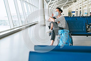 Lonely female solo traveler with backpack sitting in the empty airport passenger transfer hall in protective face mask and looking