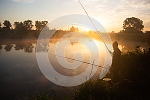 Lonely fisherman angling at foggy lake in the early morning just after golden sunrise