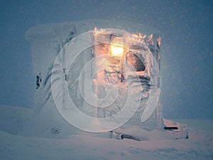 Lonely frozen and snow-covered cabin with luminous street lamp during blizzard at night