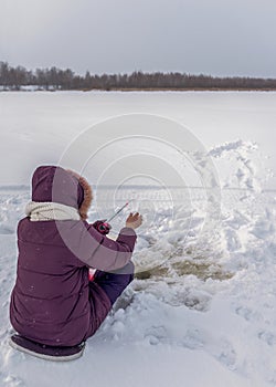 A lonely girl angler enthusiastically fishing with a winter fishing rod sitting on the ice among the flakes of white snow