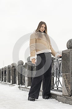 Lonely girl in beige sheepskin coat and black pants stands in snow-covered city park. Casual winter look for city