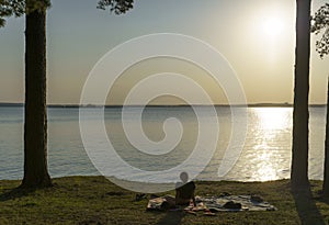 A lonely girl is resting and admiring the sunset on the shore of a forest lake. Relax, lifestyle, tranquility, rest