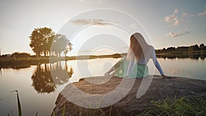 A lonely girl sits on a lake on a stone on a sunset background. Video on the move.