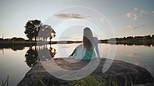 A lonely girl sits on a lake on a stone on a sunset background. Video on the move.