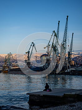 A lonely girl sits on a pier in the port. Sitting on the edge of the water. Uses a phone