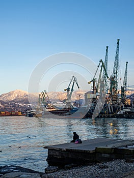 A lonely girl sits on a pier in the port. Sitting on the edge of the water. Uses a phone