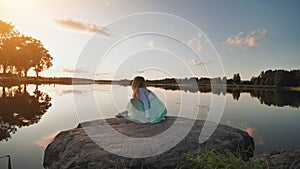 A lonely girl sits on the shore of a lake and splashes water during sunset. Video on the move.
