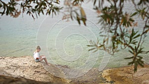 Lonely girl sitting on the beach on a rock and reading a book. Schoolgirl doing homework, summer vacation