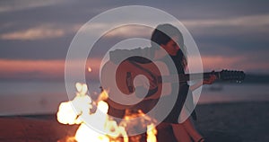 Lonely girl sitting by bonfire on the beach near forest playing acoustic guitar. Solo activity and social distancing