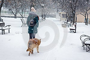Lonely girl walks in a snowy park next to a dog. A dog follows a girl during a snowfall in the park_