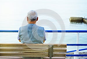 Lonely gray-haired elderly man sitting by the sea on a bench, a view from the back, contemplation of nature and a relaxing holiday
