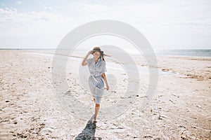 Lonely happy young woman in a hat and dress walks on a deserted sea beach on a sunny day. Summer vacation