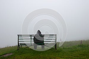 a lonely hooded figure, back to camera. Sitting on a bench in the countryside. On a cloudy rainy day