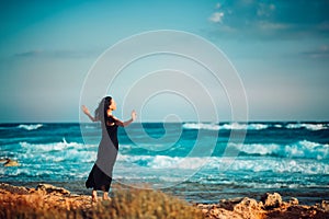 Lonely inspired woman in black dress staying on seaside dreaming and looking to sea on windy sunset day. Back view. Emotions