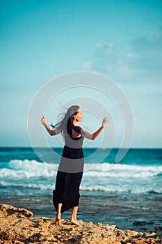 Lonely inspired woman in black dress staying on seaside dreaming and looking to sea on windy sunset day. Back view. Emotions