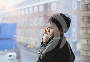 Lonely Kid with sad face hugging teddy bear standing next to window, Bored Child holding soft toy standing alone looking through