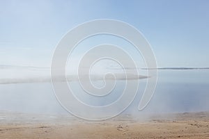 Lonely lifeless lake shore in early morning with soft haze above water and yellow sand coast with cape in fog. Mysterious.