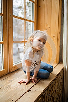 Lonely Little Blonde Girl Sitting on a wooden windowsill Near Big Window In New House.