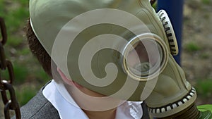 Lonely little boy in white school shirt sit on a swing with metal rusty chains in military protective gas mask on playground